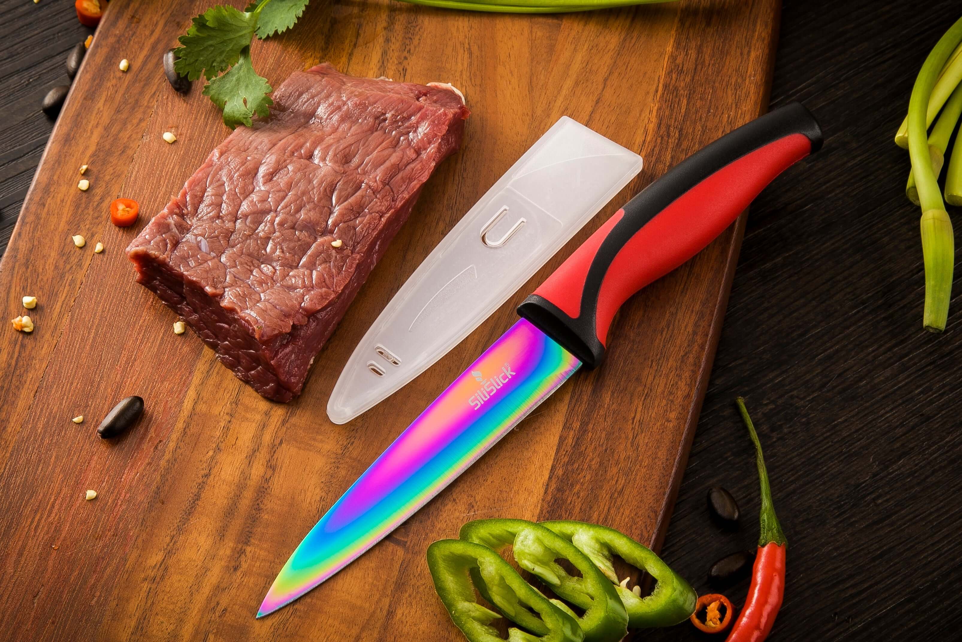 Knife with a colorful blade on a wooden cutting board with raw meat and vegetables.