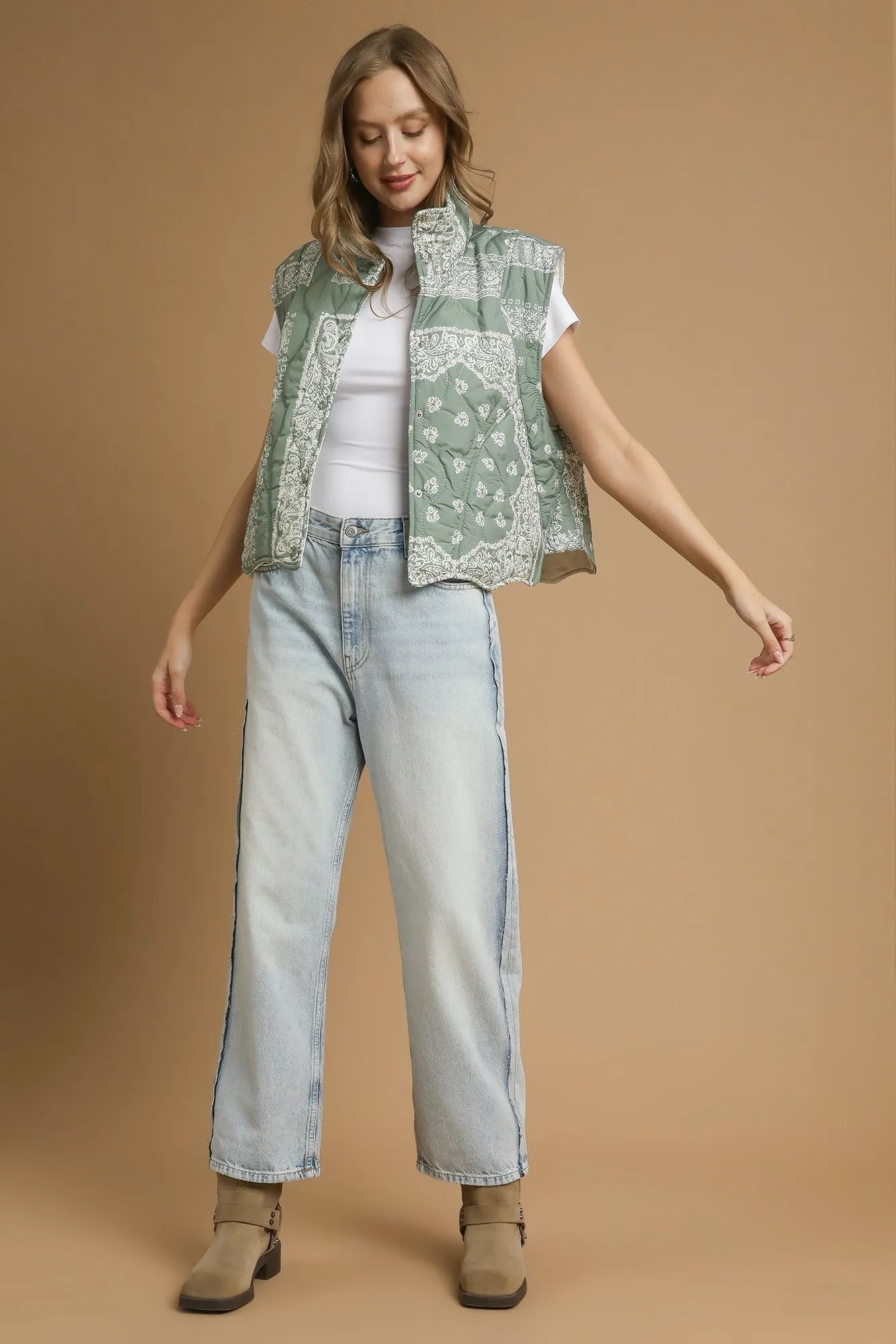 Woman wearing an olive green, bandana-patterned vest over a white shirt and light blue jeans with tan colored boots on a brown background