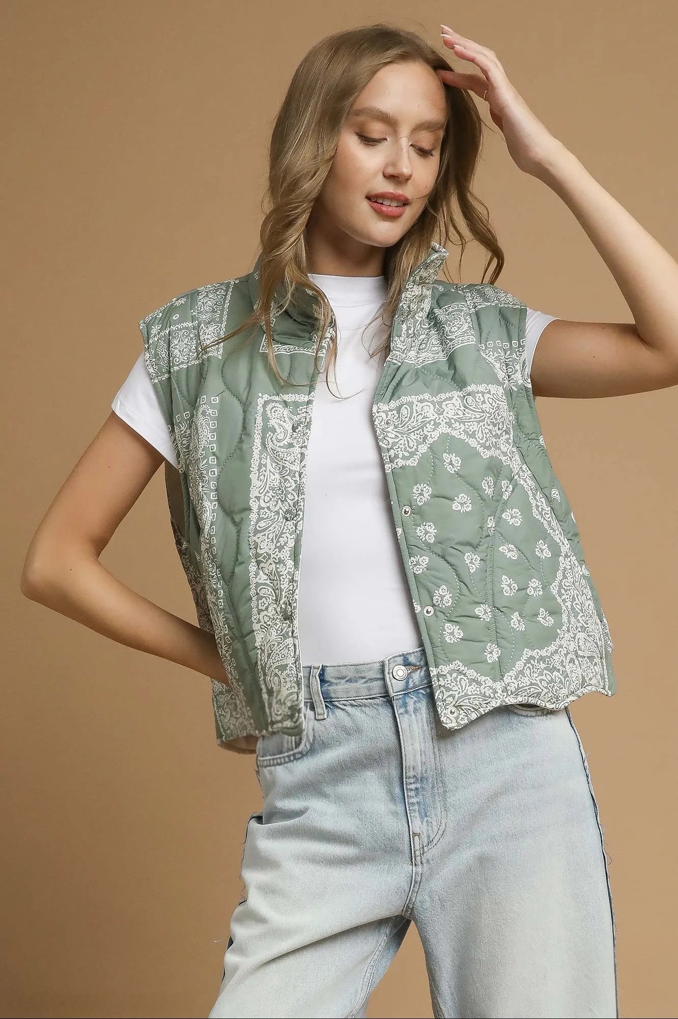 Woman wearing an olive green, bandana-patterned vest over a white shirt and light blue jeans on a brown background